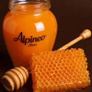 A jar of pale Acacia honey with a wooden honey dipper resting on the lid and a bloom in the background.